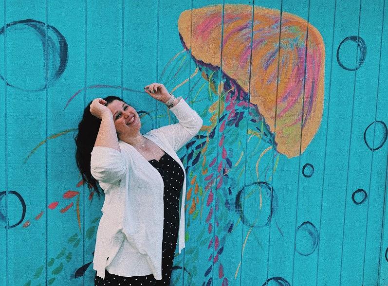 Dannie Johnson, a woman, is standing in front of a teal wall. There is a colorful jellyfish painting on the wall surrounded by painted bubbles.